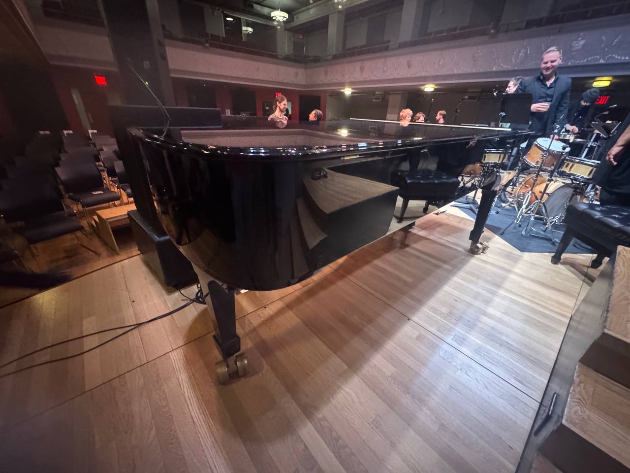 Black grand piano beside a drum kit in a performance hall